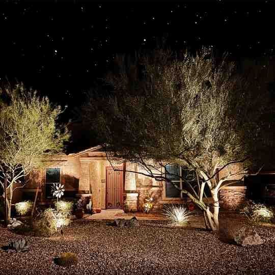 Desert house at night with warm exterior lights, trees, and a gravel yard