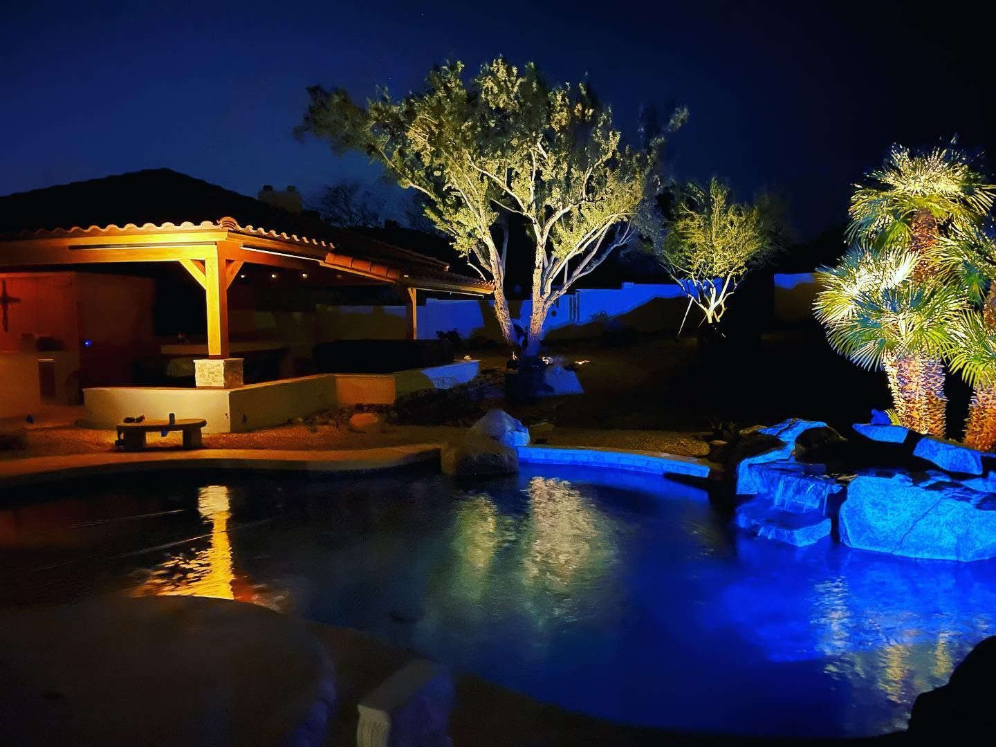 Lit poolside patio at night with illuminated trees and blue water reflections
