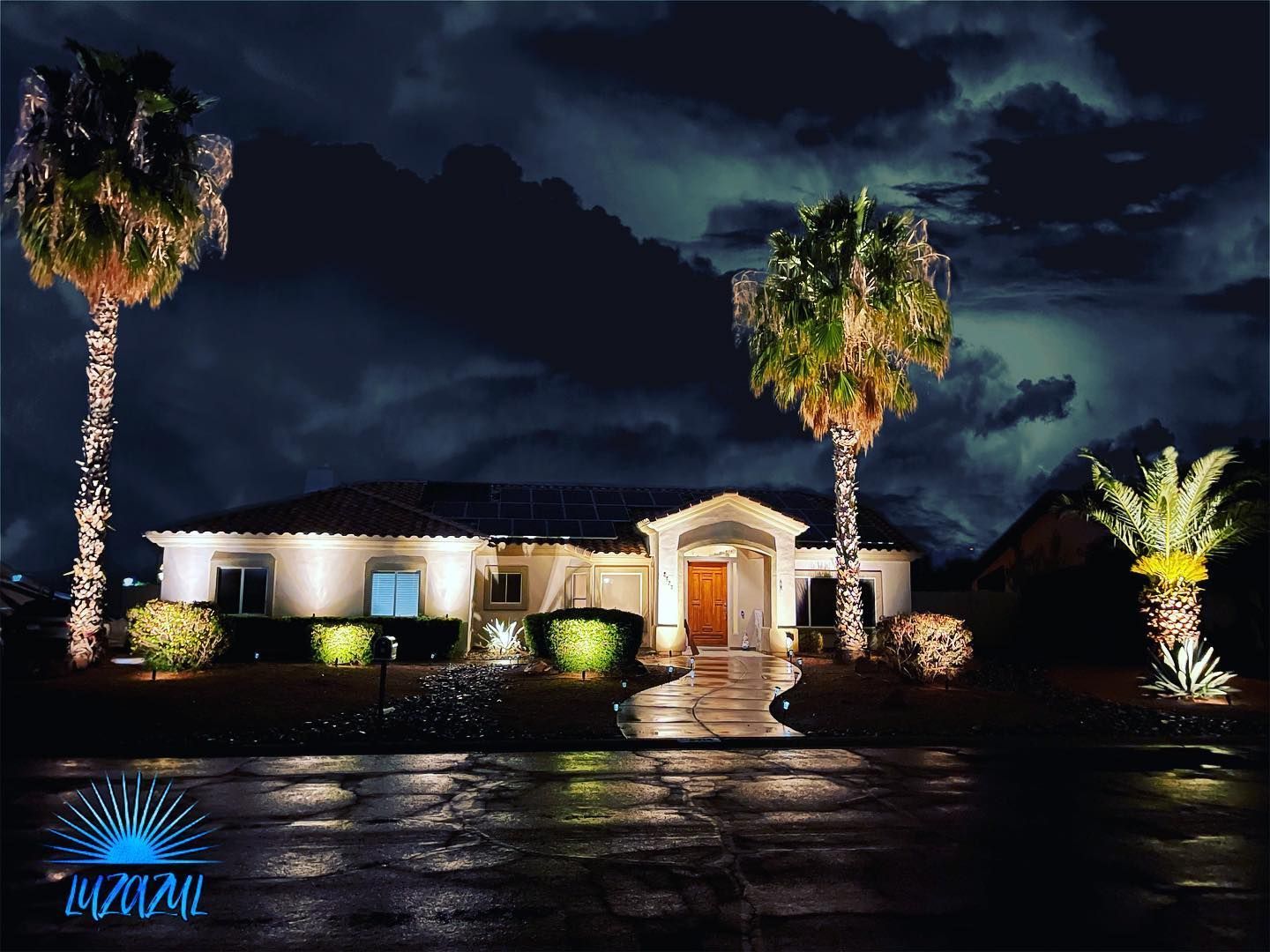 Single-story house lit at night, with palm trees and wet driveway under a dark cloudy sky