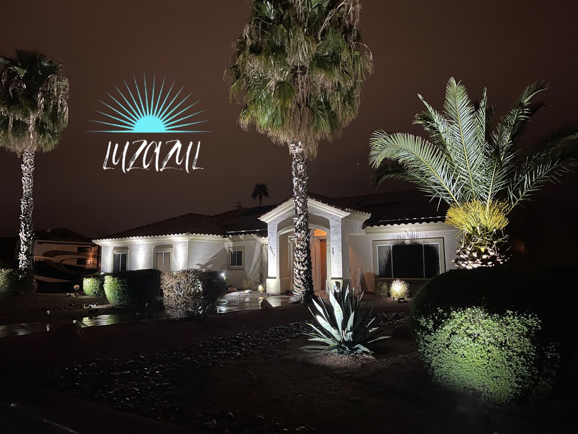 Nighttime view of a lit modern house with palm trees and a glowing sign in the front yard