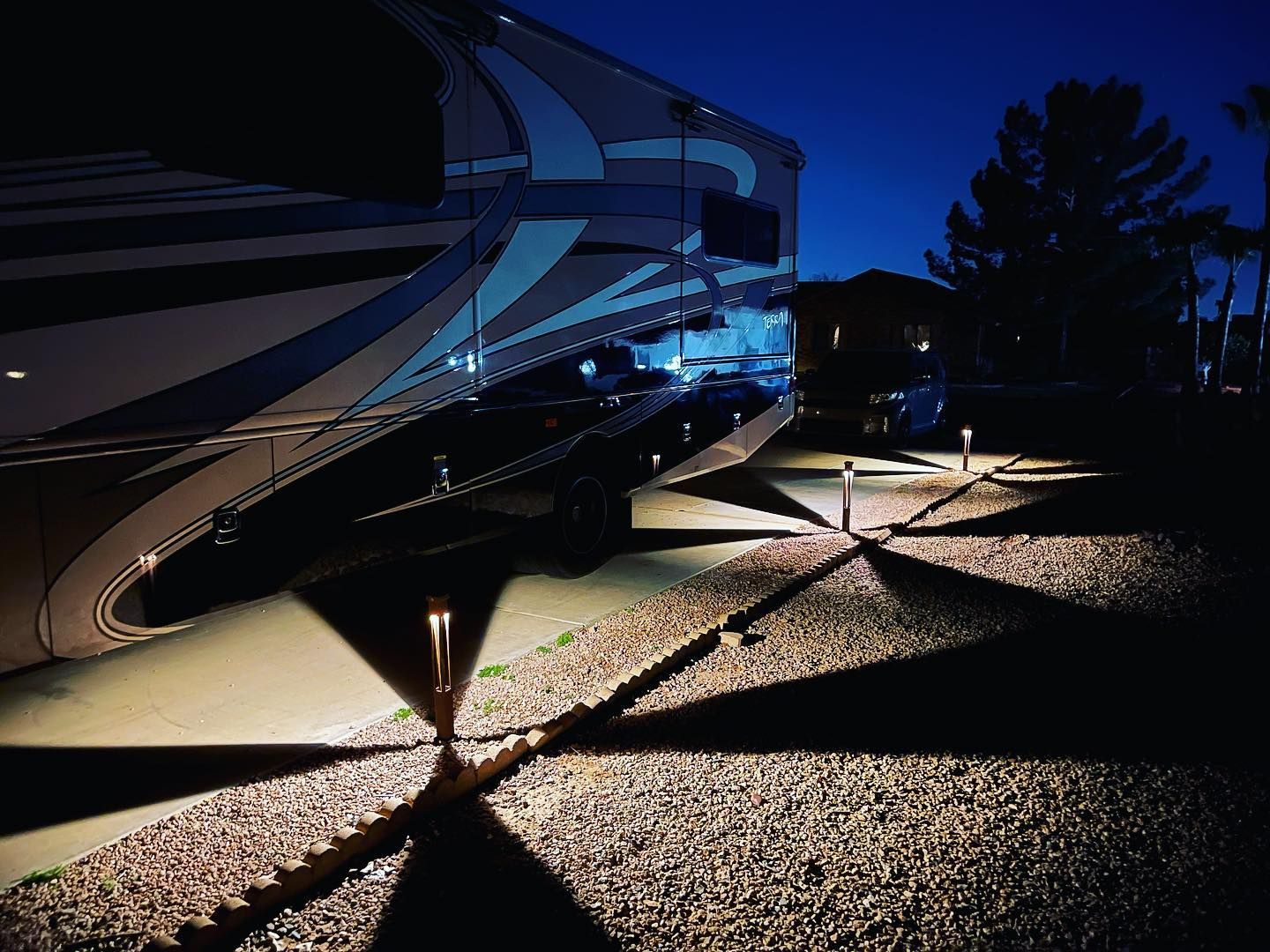 Nighttime RV parked by a gravel path, lit by blue and warm lights near a building.