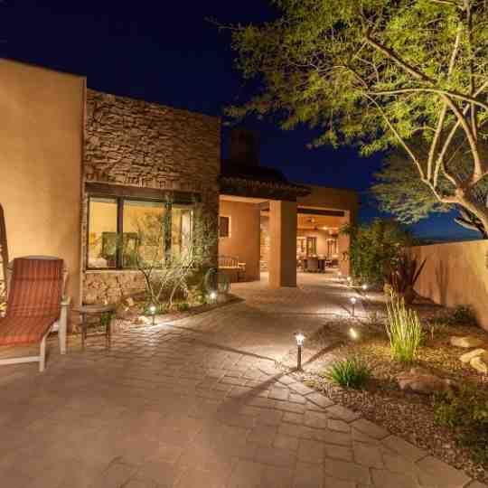 Warmly lit modern house exterior at night with stone walkway, desert landscaping, and a tree in the foreground