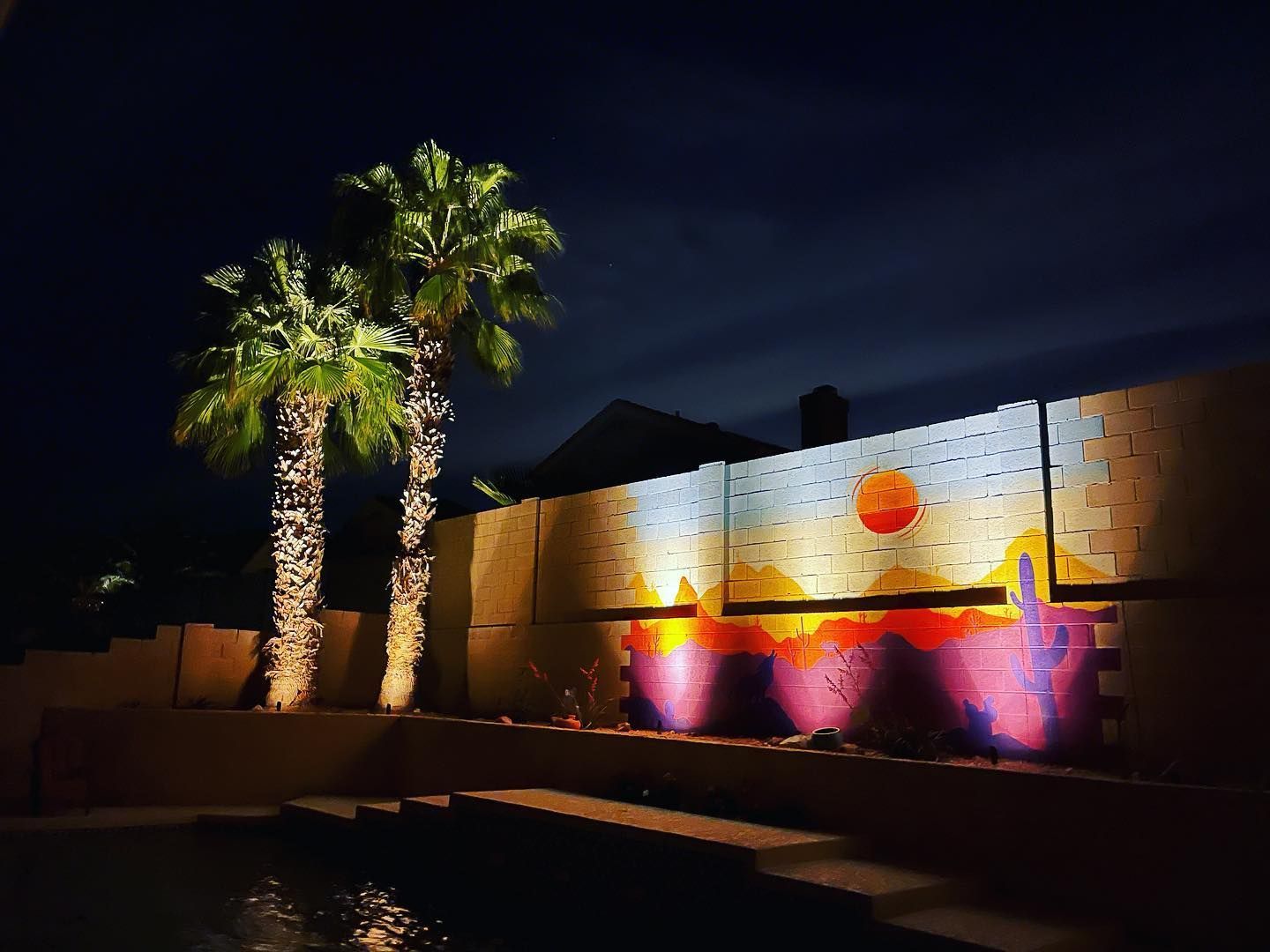 Night-lit wall mural with palm trees and colorful purple-pink uplighting in a courtyard