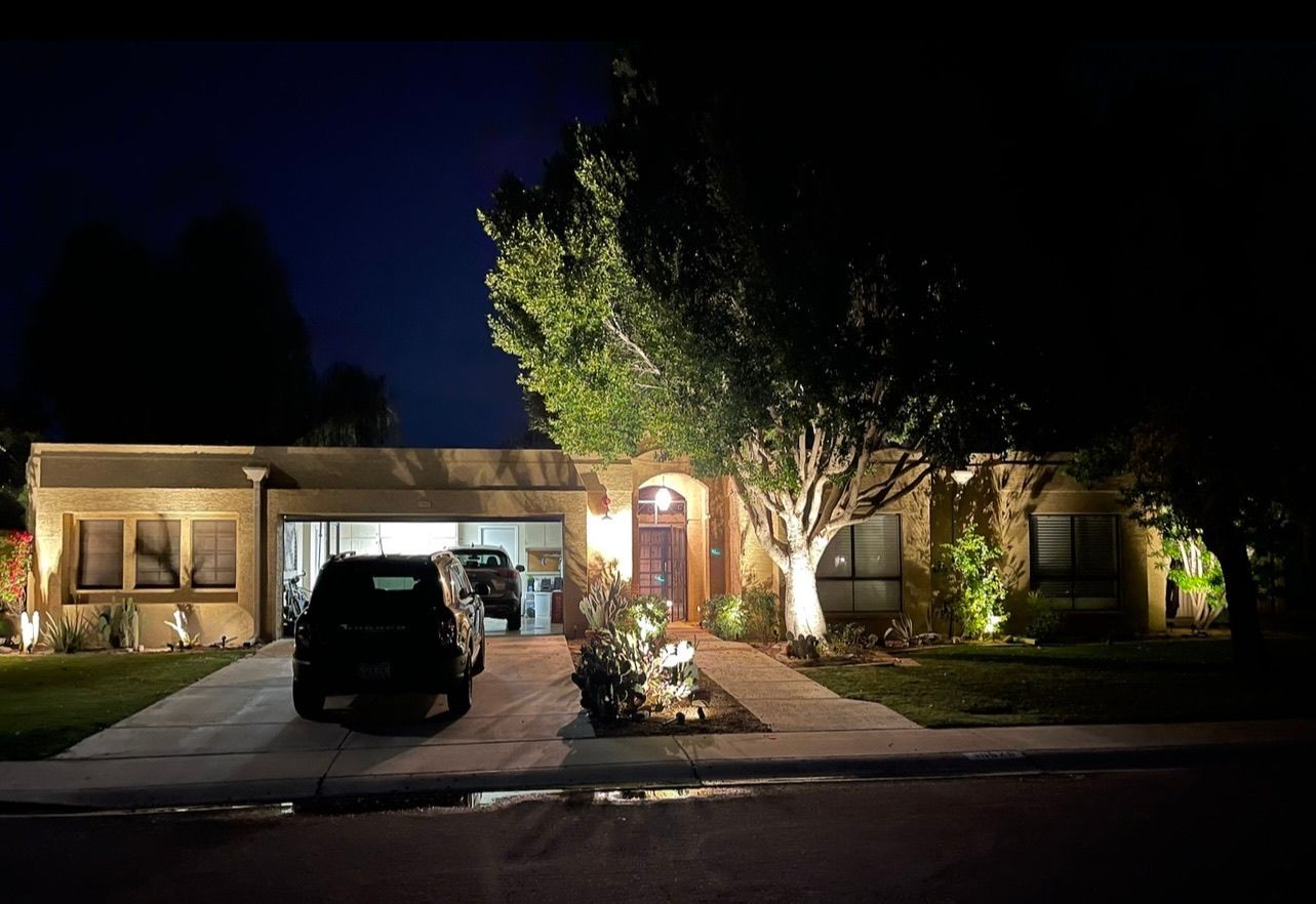 Nighttime view of a lit suburban house with a car parked in the driveway and a large tree in front.