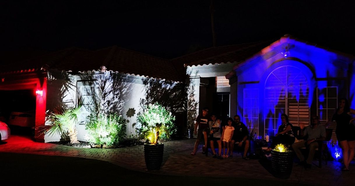 House exterior at night lit with red, white, and blue holiday lights, with small trees and potted plants in front.