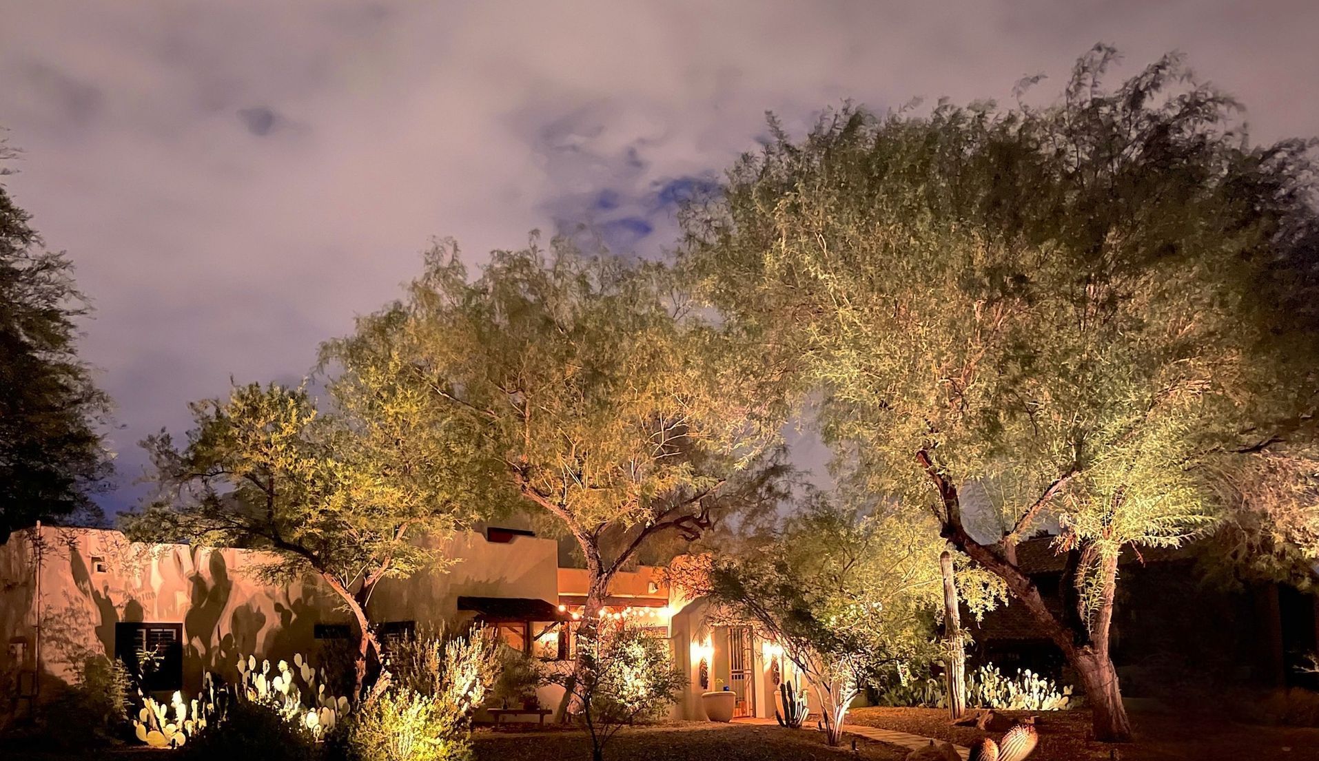 Nighttime desert home lit by warm lights, with tall trees and a cloudy sky.
