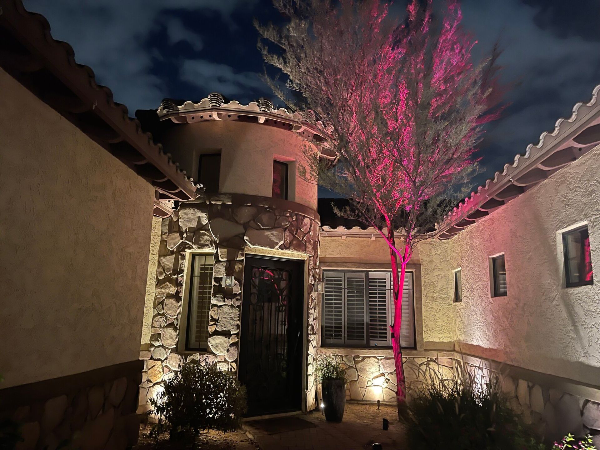 Nighttime courtyard with a stone house, lit doorway, and pink-lit tree under a dark sky