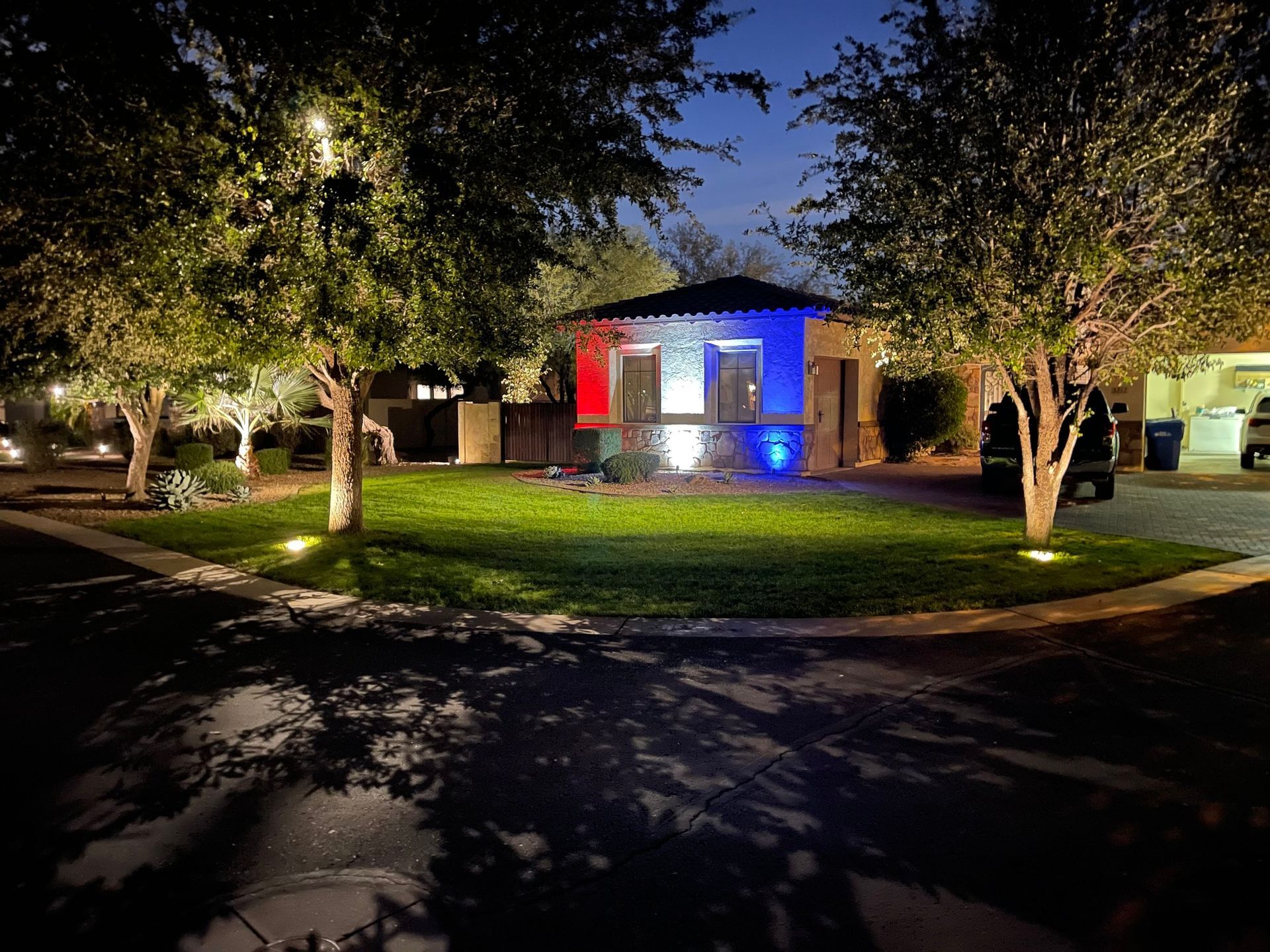 Nighttime view of a small lit building with colorful lights, surrounded by trees and a grassy lawn.