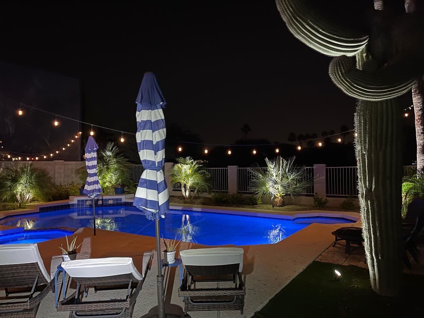 Nighttime poolside patio with lounge chairs, striped umbrellas, string lights, and cacti in the background
