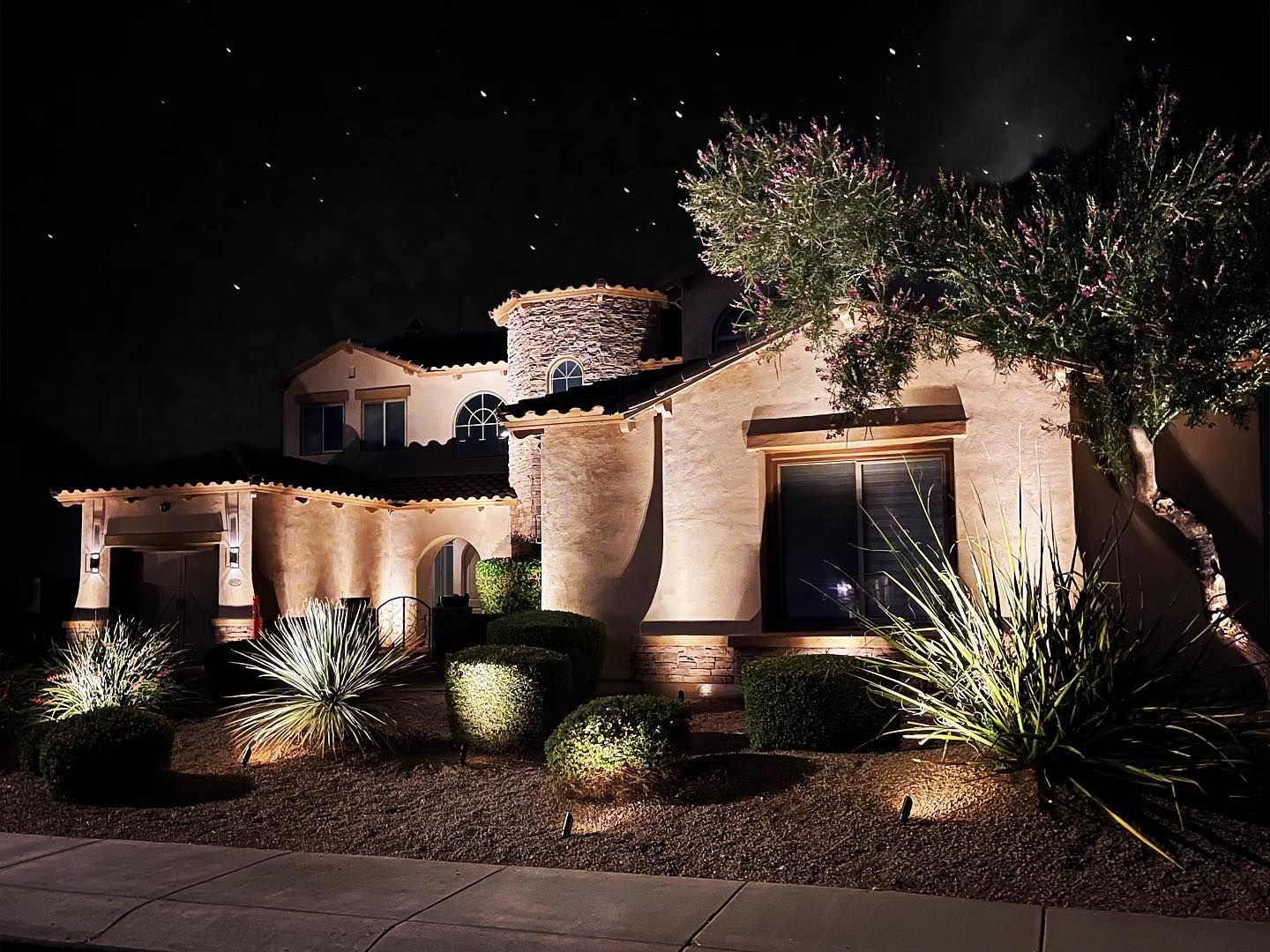 Illuminated stucco house at night with desert landscaping and trees in front