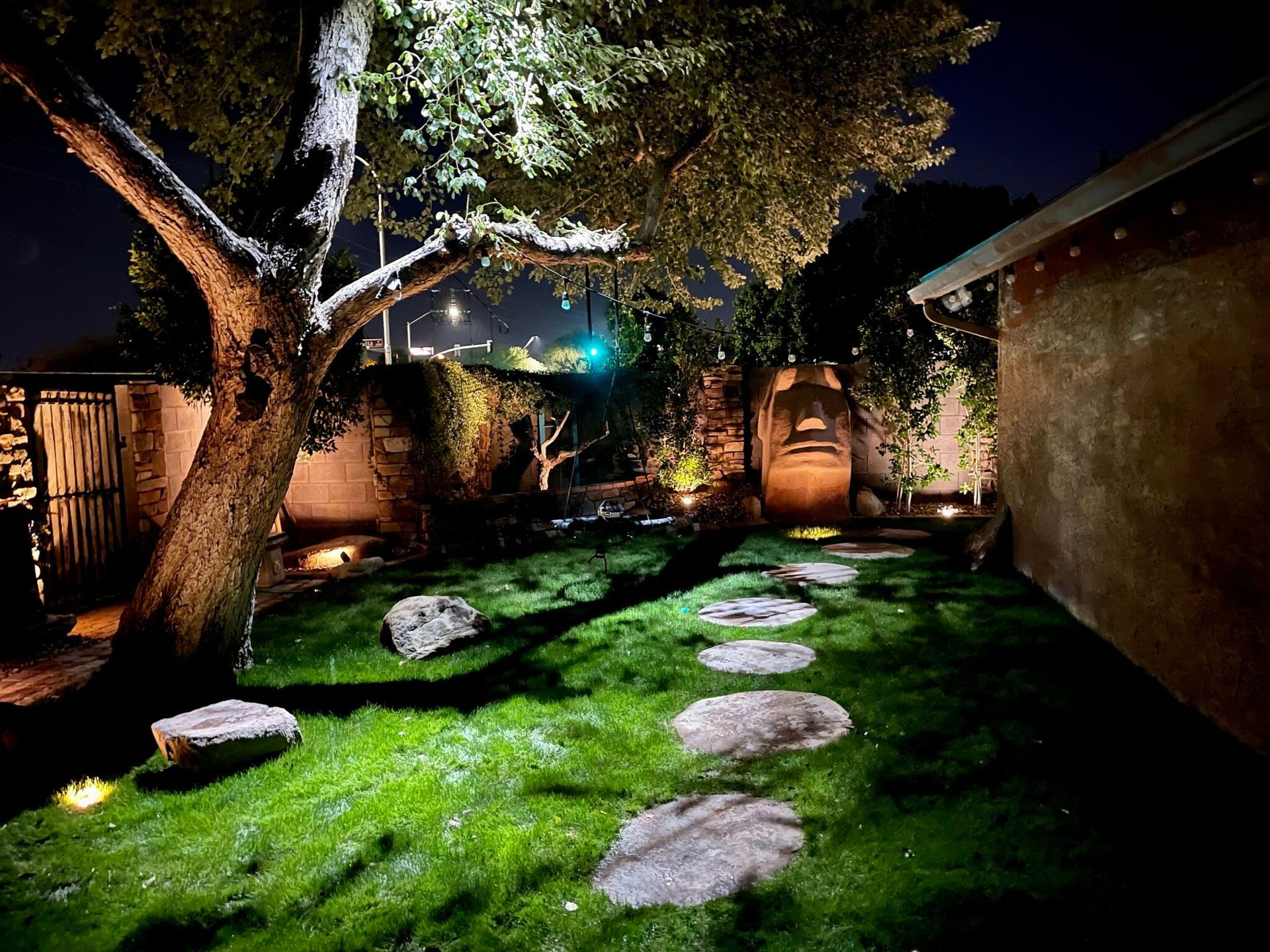 Illuminated garden path with stepping stones, lush grass, trees, and brick walls at night