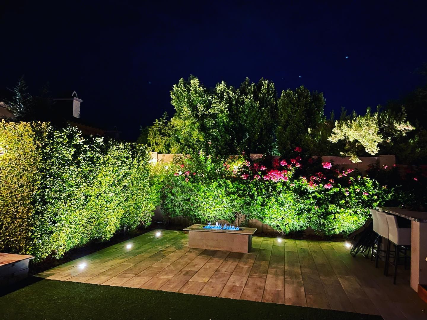 Lit backyard patio with wooden deck, bright garden lights, and lush green shrubs under a dark sky