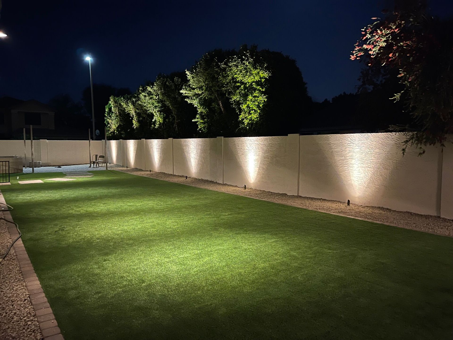 Lit backyard lawn at night with spotlights, white wall, and trees lining the edge.
