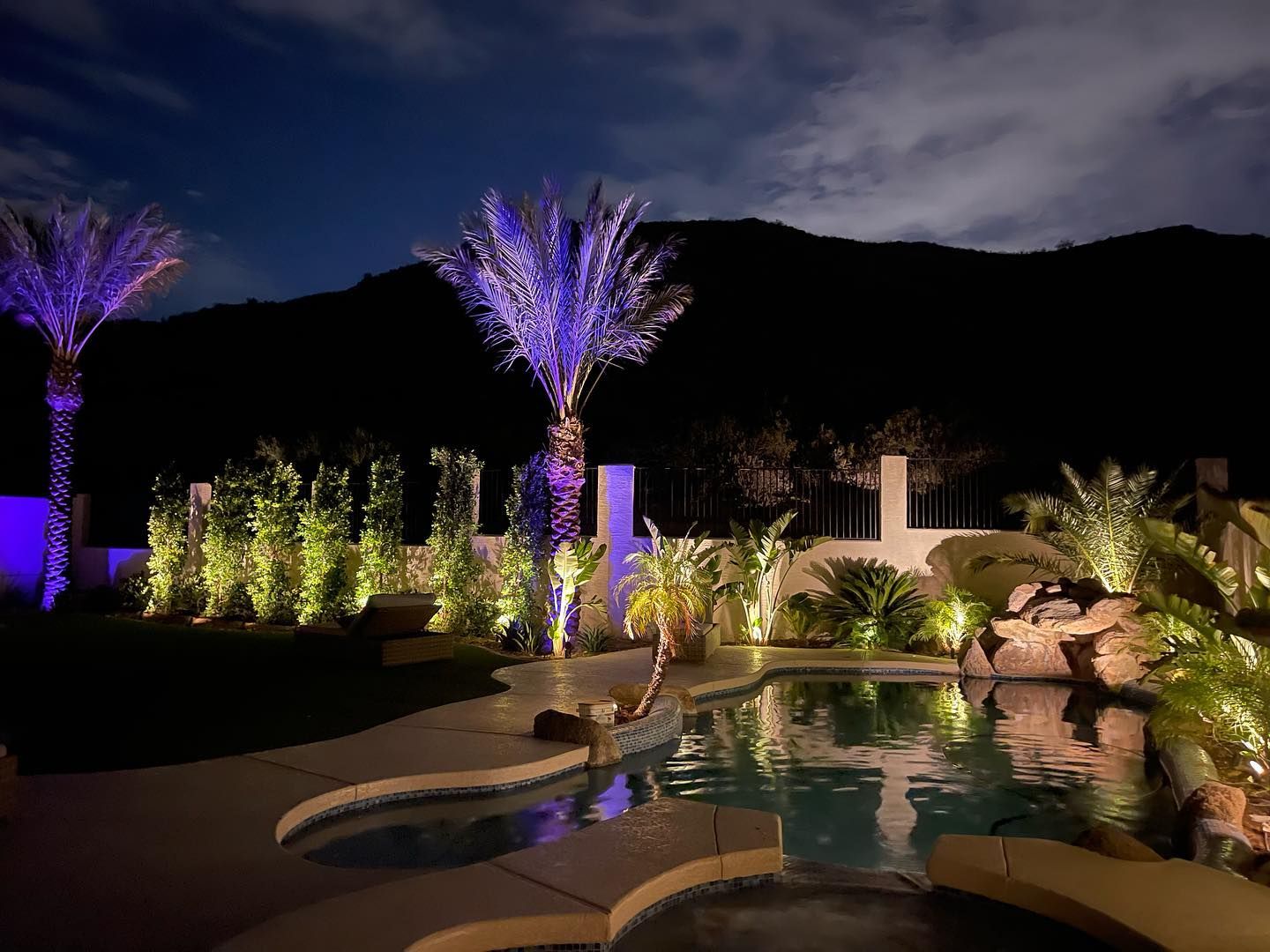 Nighttime resort pool with glowing purple palm trees, rocks, and a waterfall against mountains