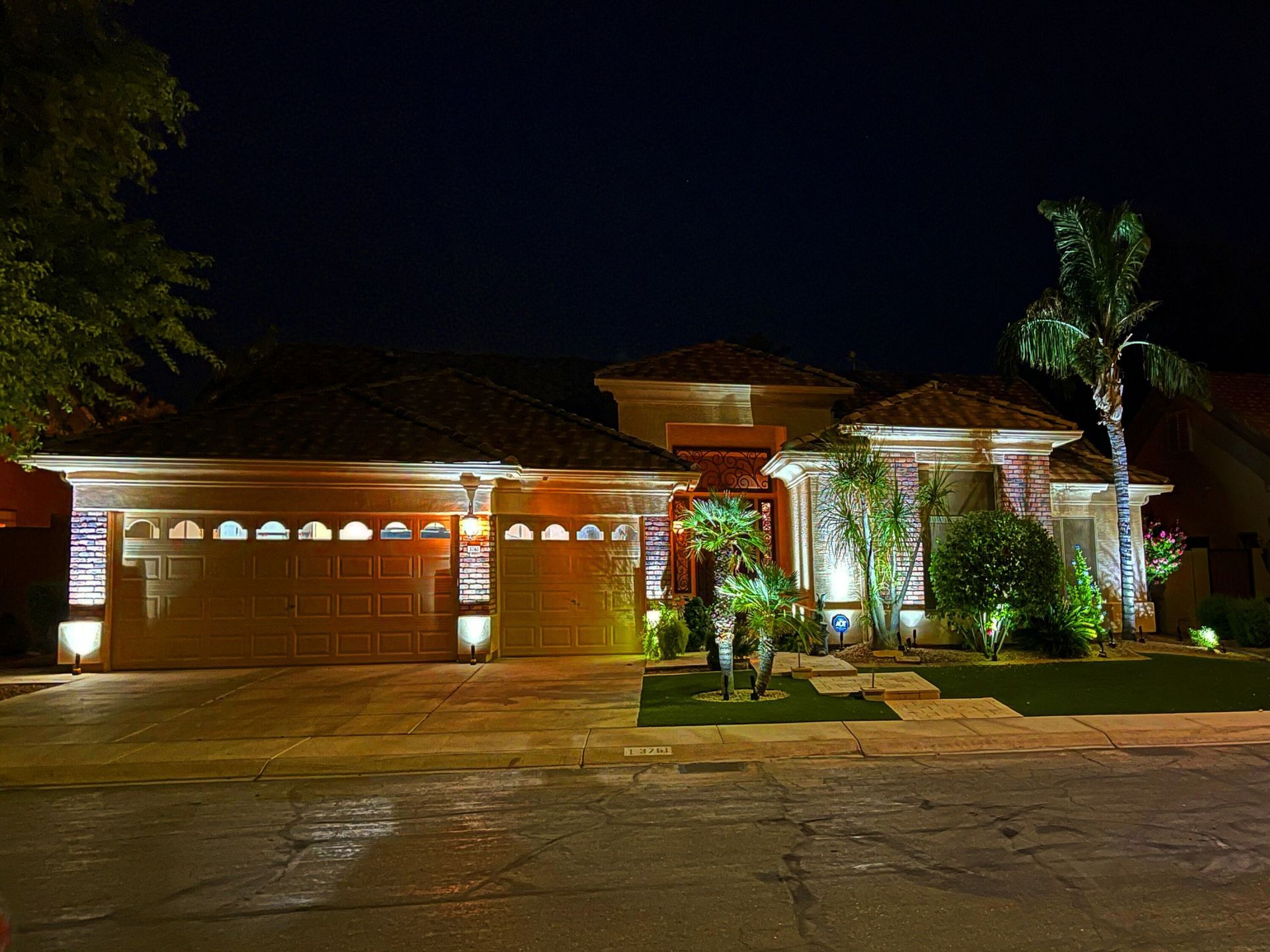 Lit suburban house at night with illuminated garage, palm trees, and landscaped front yard