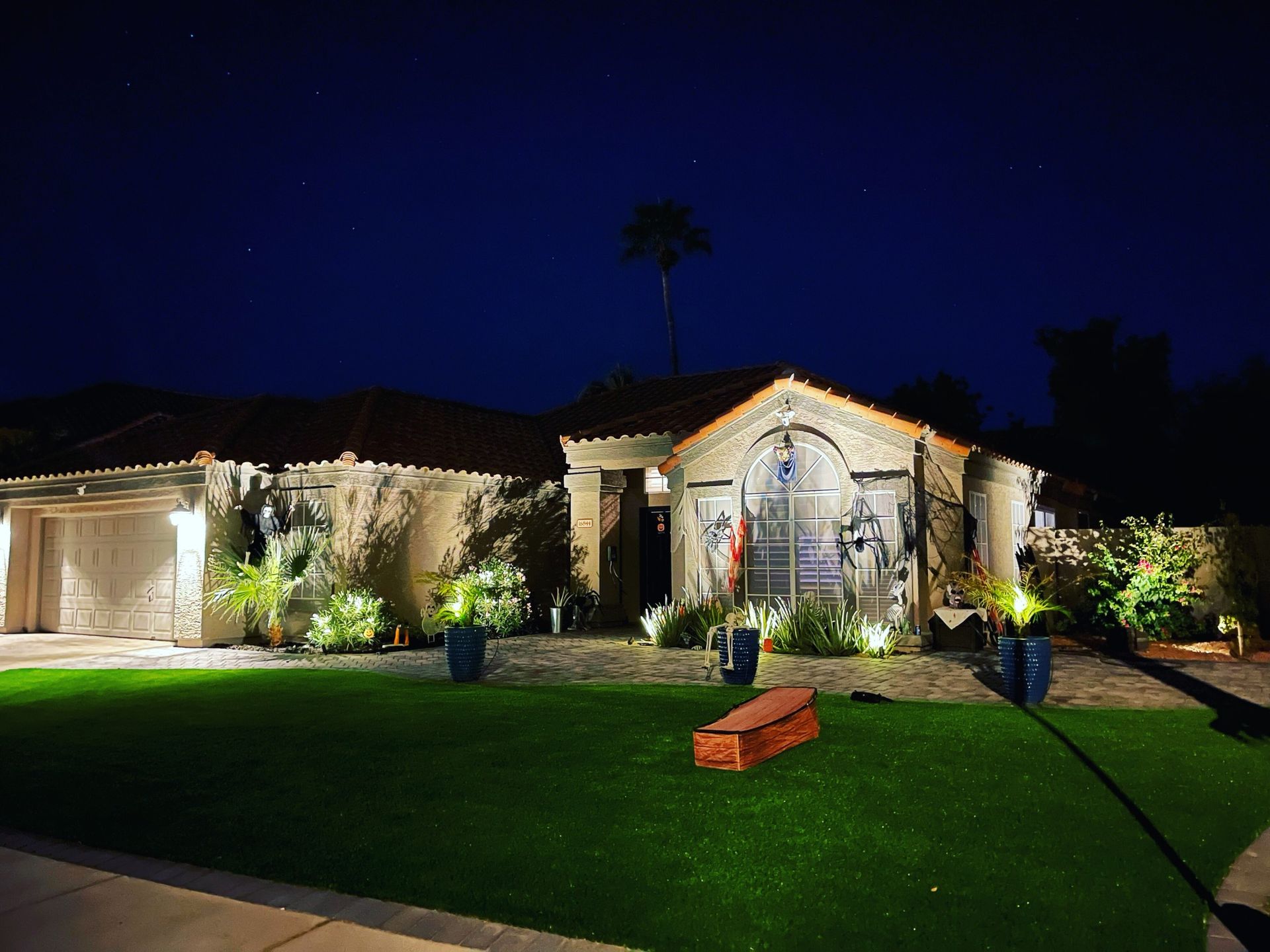 Well-lit suburban house at night with a green lawn and palm tree silhouettes under a dark blue sky