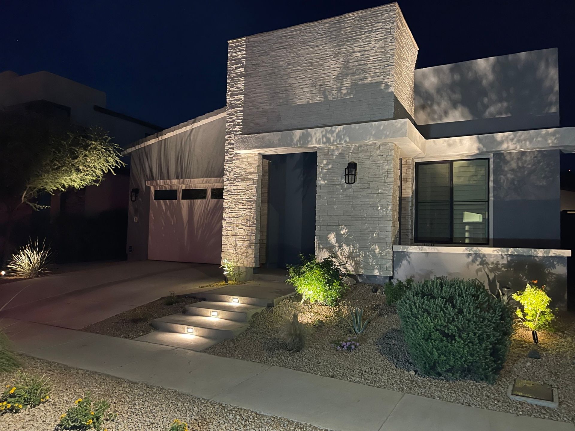 Modern house exterior at night with stone facade, lit walkway, and desert landscaping