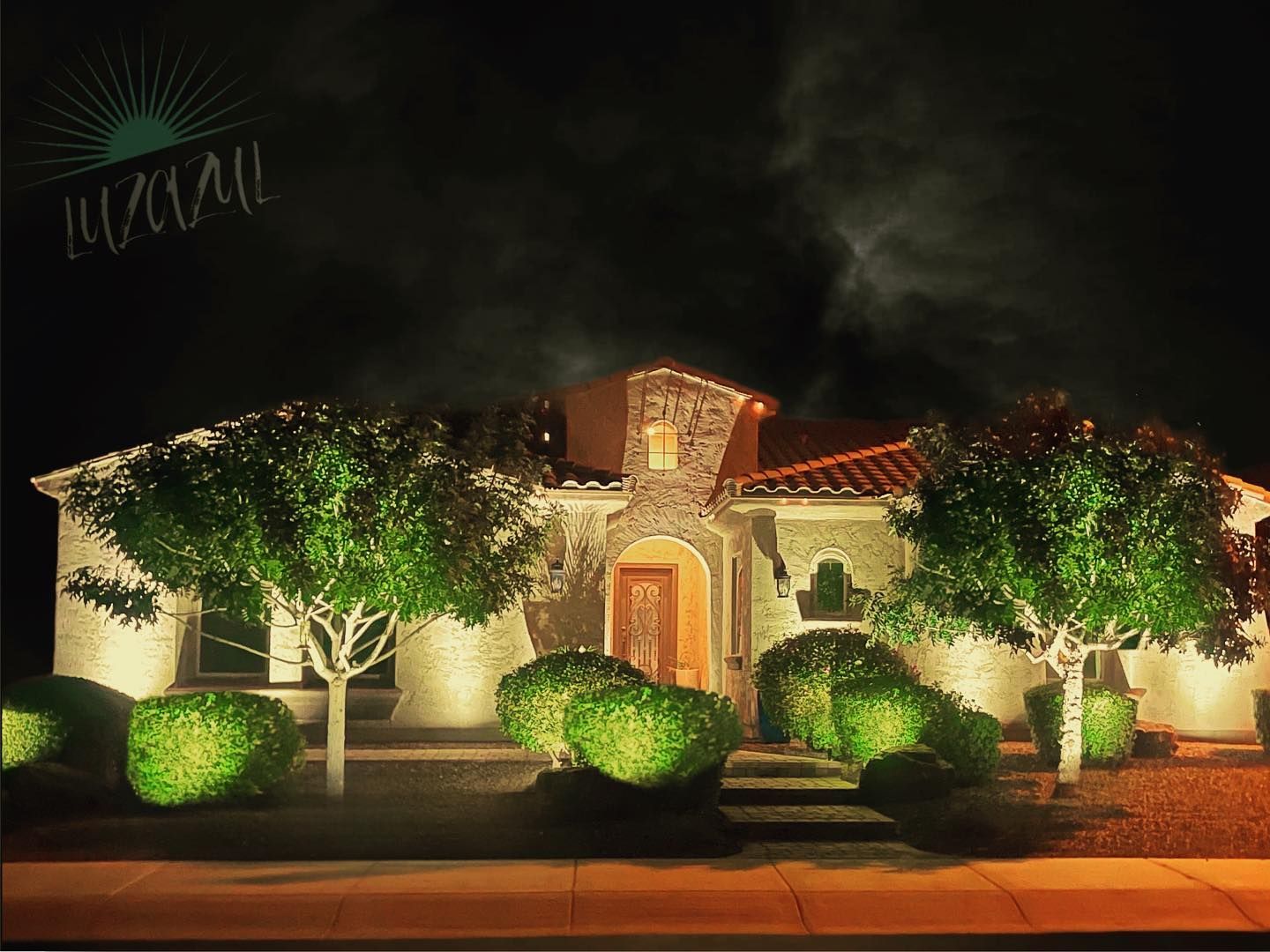 Well-lit stucco house with red tile roof and landscaped front yard at night