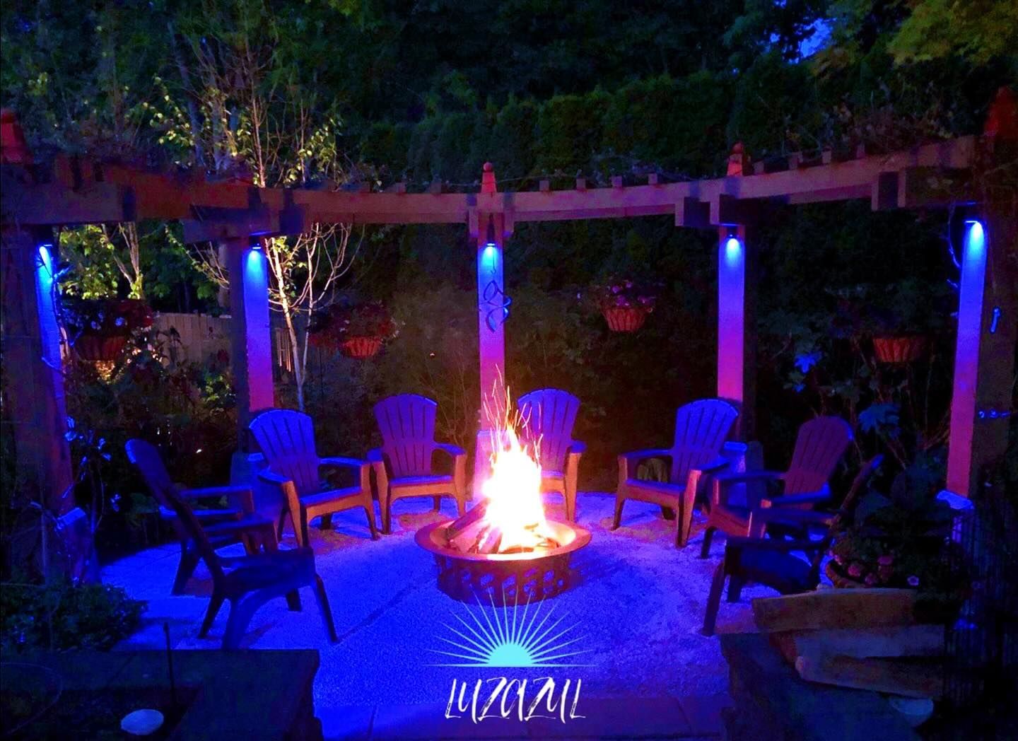 Outdoor fire pit surrounded by blue-lit chairs and plants at night