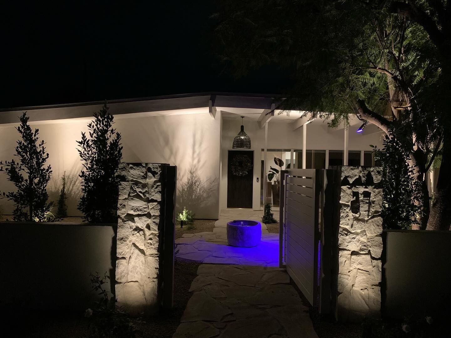 Nighttime view of a lit house entrance with stone pillars, trees, and blue uplighting on the walkway