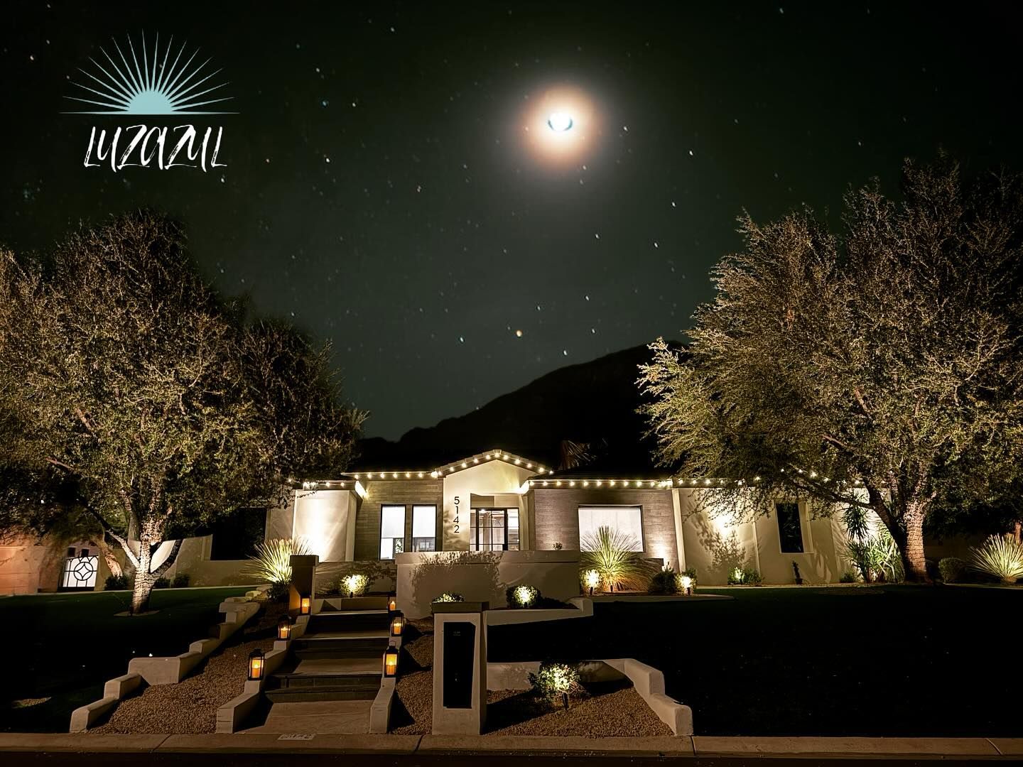 Nighttime view of a lit stone mansion under the moon, framed by trees and a landscaped entrance.