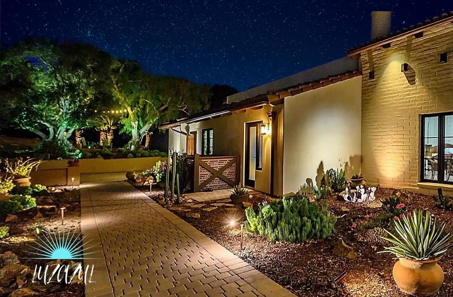 Nighttime view of a warmly lit house patio with brick walkway, garden plants, and trees