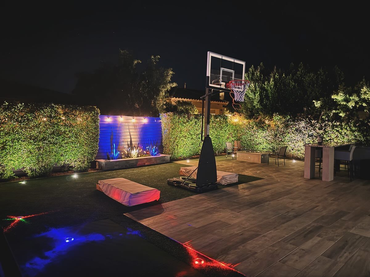 Nighttime patio with a basketball hoop, lit fountain, and colorful ground lights beside hedges