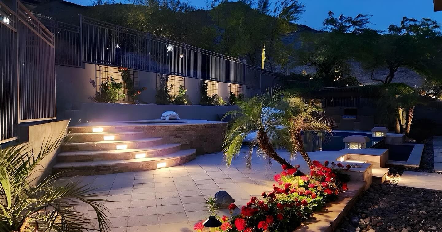 Illuminated patio steps and seating area at dusk, with palm trees, flowers, and warm garden lights.