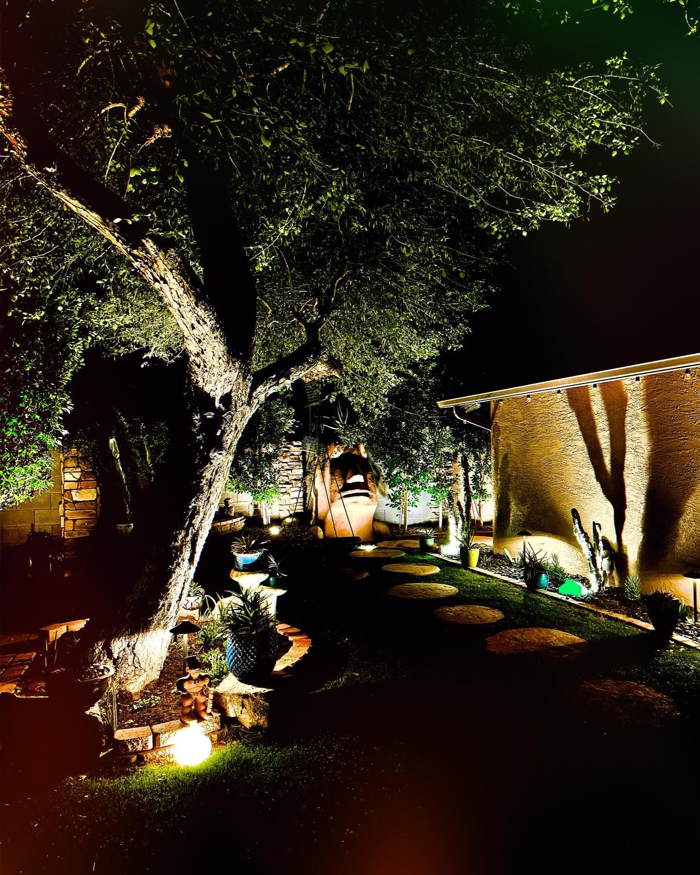 Nighttime garden path lit by warm ground lights, with trees and a stone wall