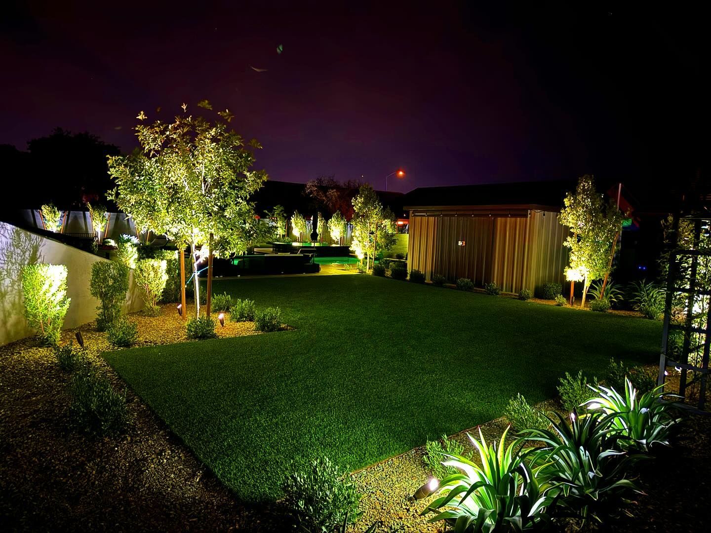 Illuminated backyard lawn with trees, shrubs, and a lit shed at night under a dark sky