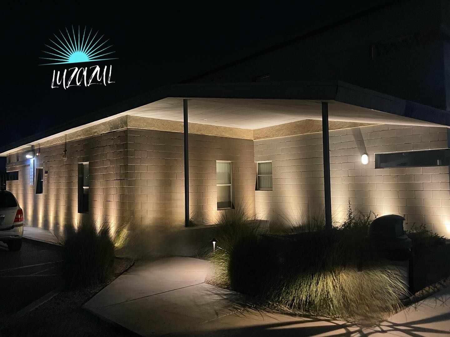 Night-lit modern brick building entrance with warm wall lights and a glowing sign above the corner.