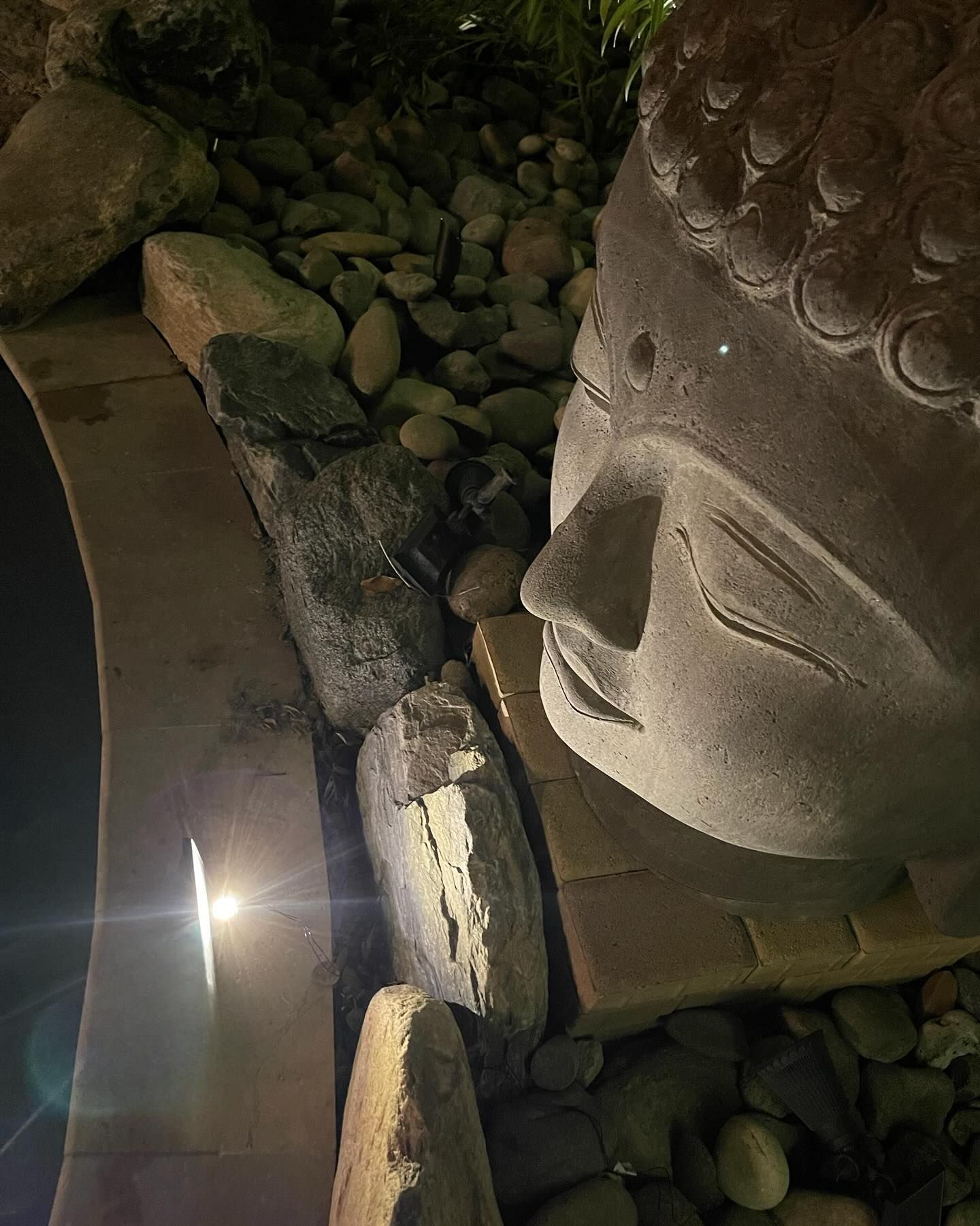 Large stone Buddha face beside a small water feature and mossy rocks in a dim grotto