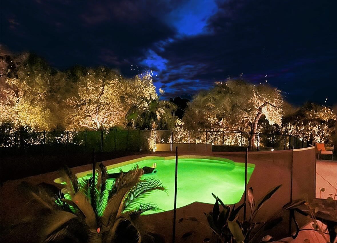 Nighttime courtyard with glowing green pool, lit trees, and a dark blue sky