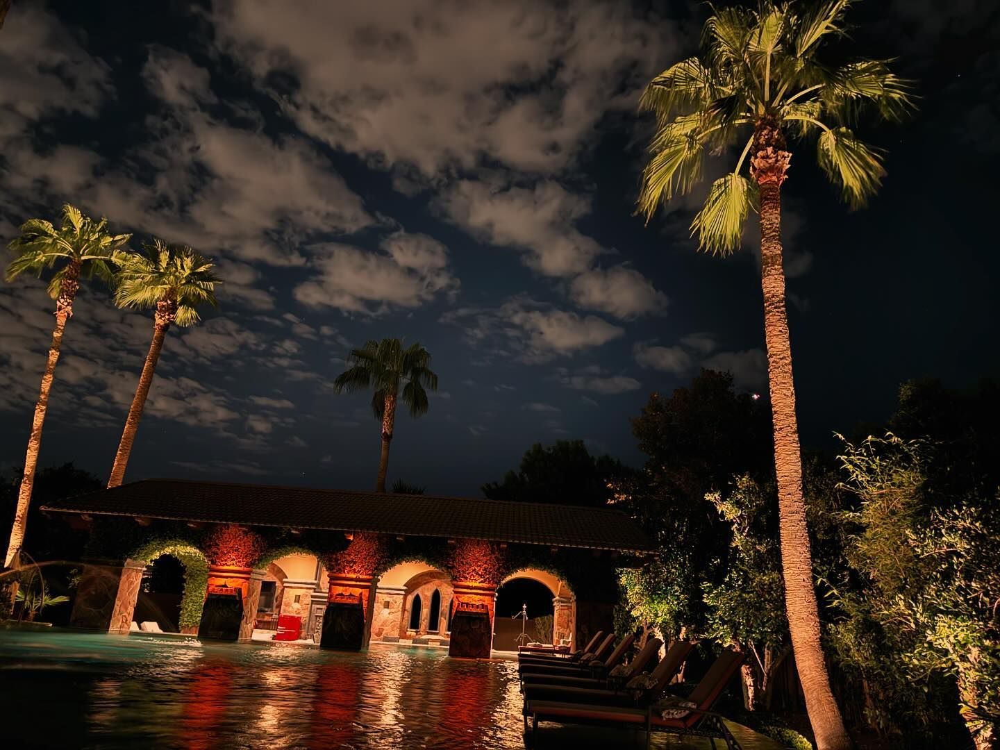 Nighttime tropical courtyard with palm trees and a warmly lit arched building reflecting in water