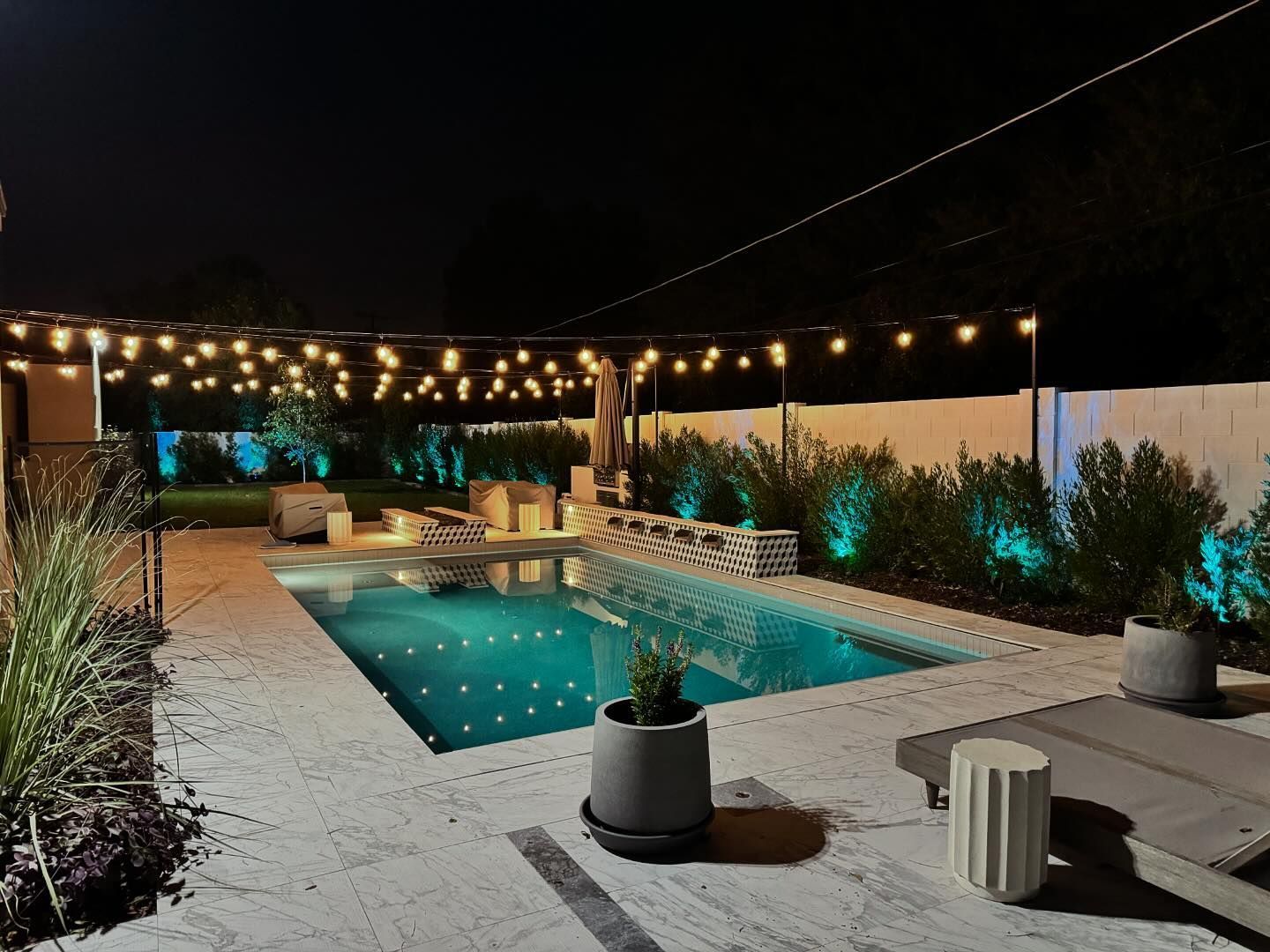 Backyard pool at night with string lights, lounge chairs, and potted plants