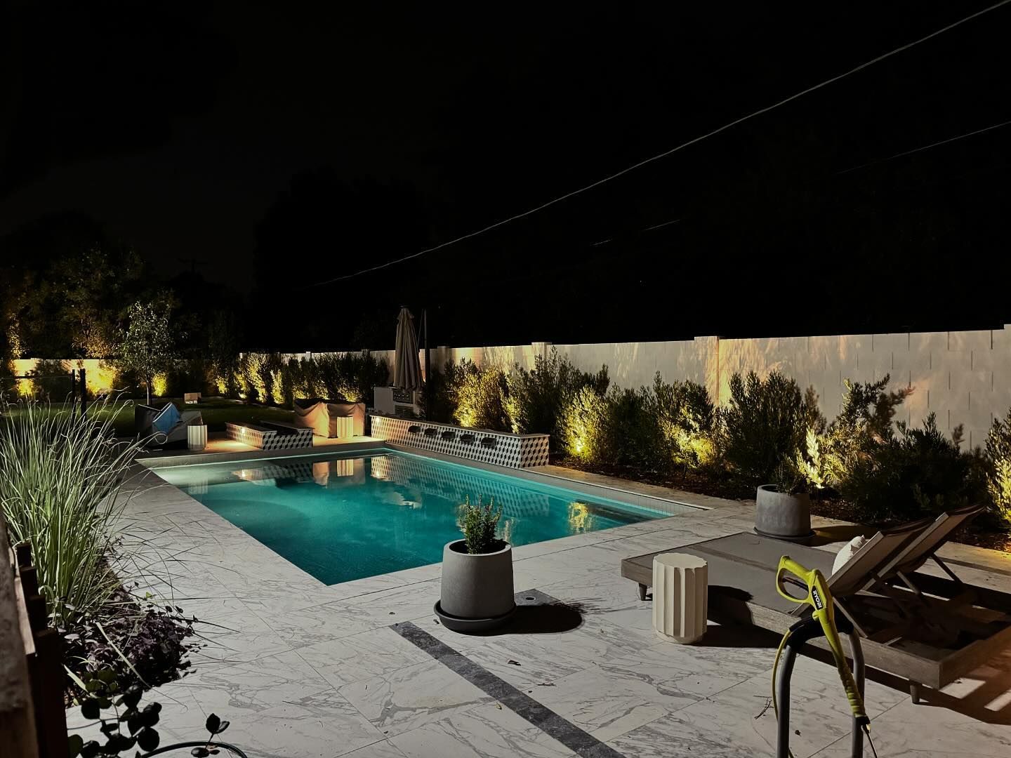 Nighttime backyard pool with lounge chairs, potted plants, and string lights along a wall