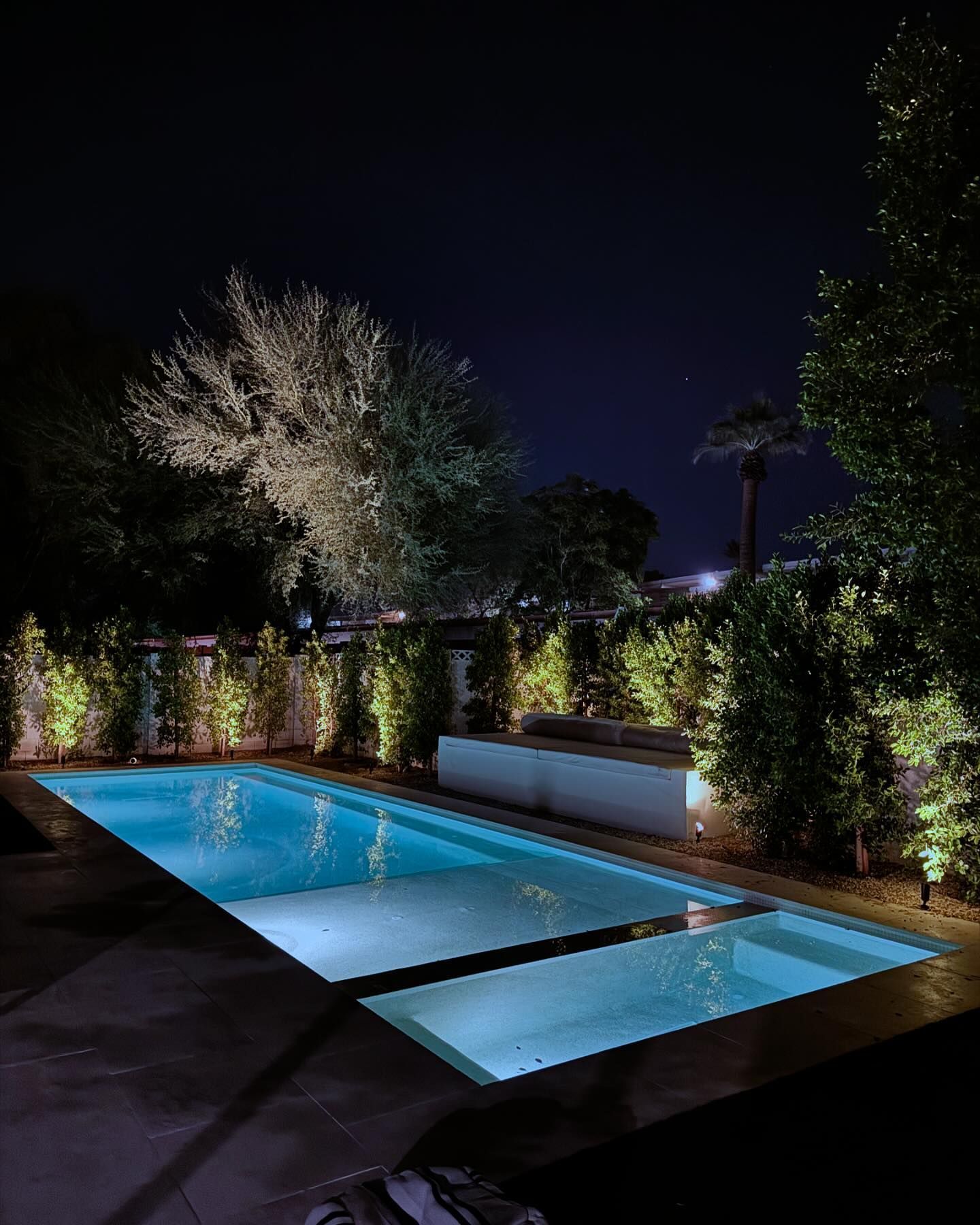 Modern backyard pool at night with blue-lit water and lush landscaping illuminating the deck