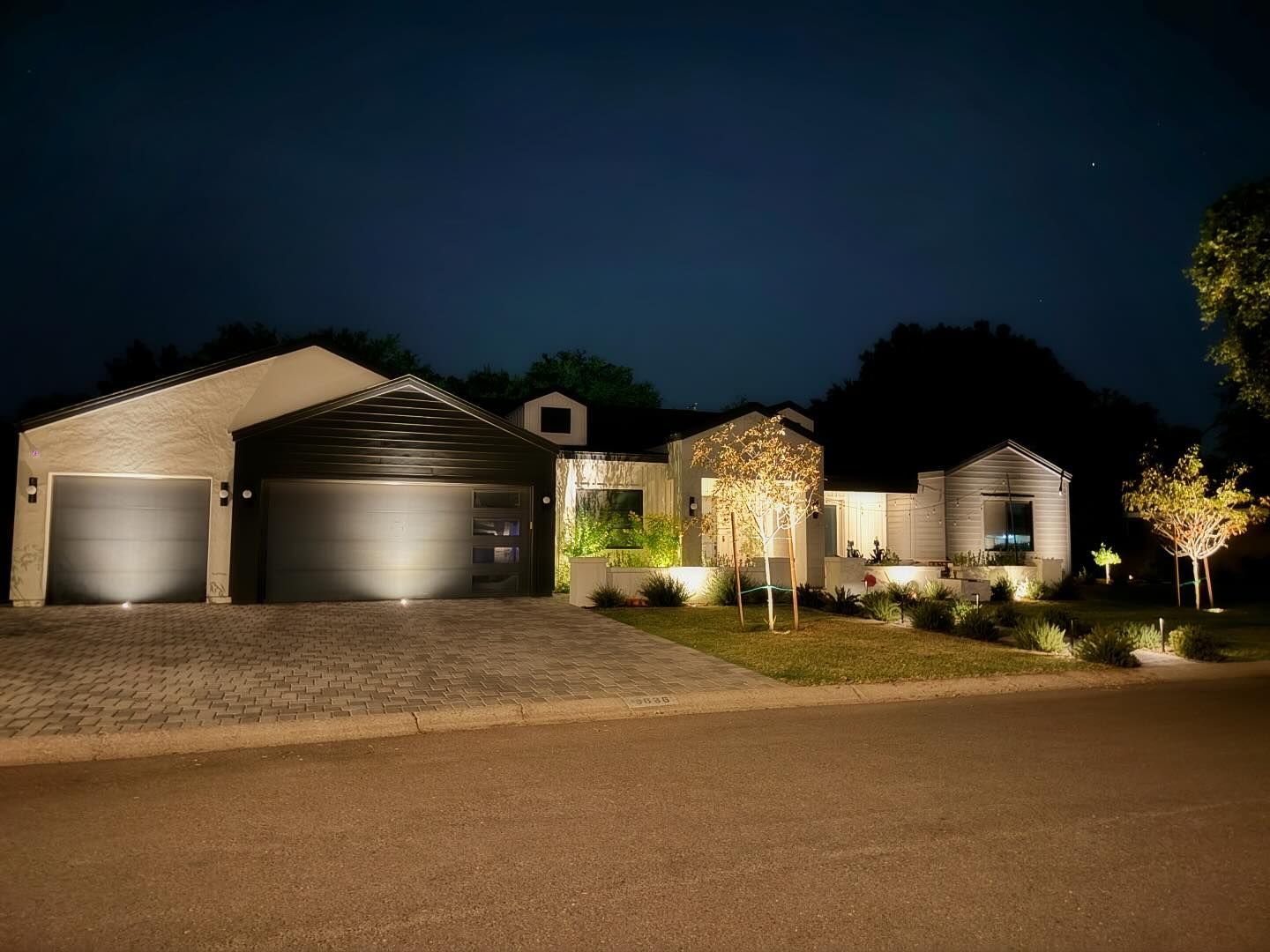 Well-lit suburban house at night with two garages, front yard lights, and a quiet street