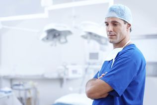 Surgeon in blue scrubs, arms crossed, in a bright operating room, holding surgical mask.