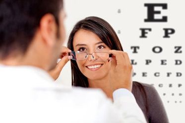 Optometrist fitting eyeglasses on a smiling woman in front of an eye chart.