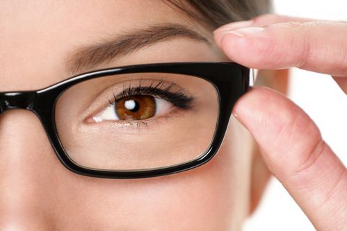 Woman's eye with brown iris, framed by black glasses, hand adjusting glasses.