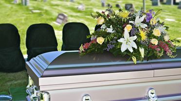 A casket with floral arrangement at an outdoor funeral, with black chairs and a grassy background.