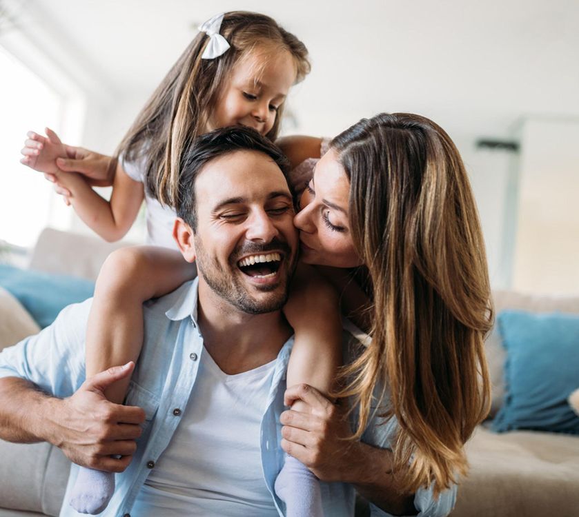 Family laughing together, daughter on father's shoulders, mother kissing him.
