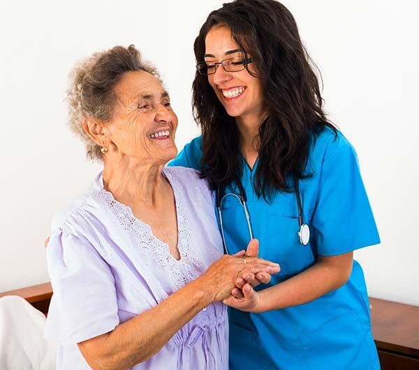 Smiling healthcare worker in blue scrubs supports elderly person in lavender clothing.