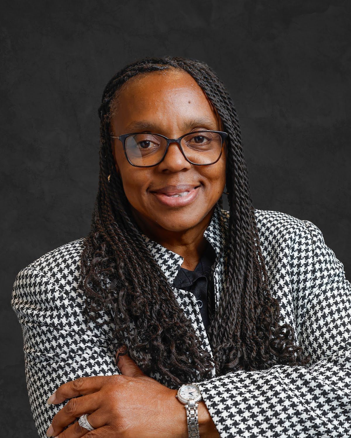 Woman with glasses, braids, and a black-and-white patterned jacket, arms crossed.