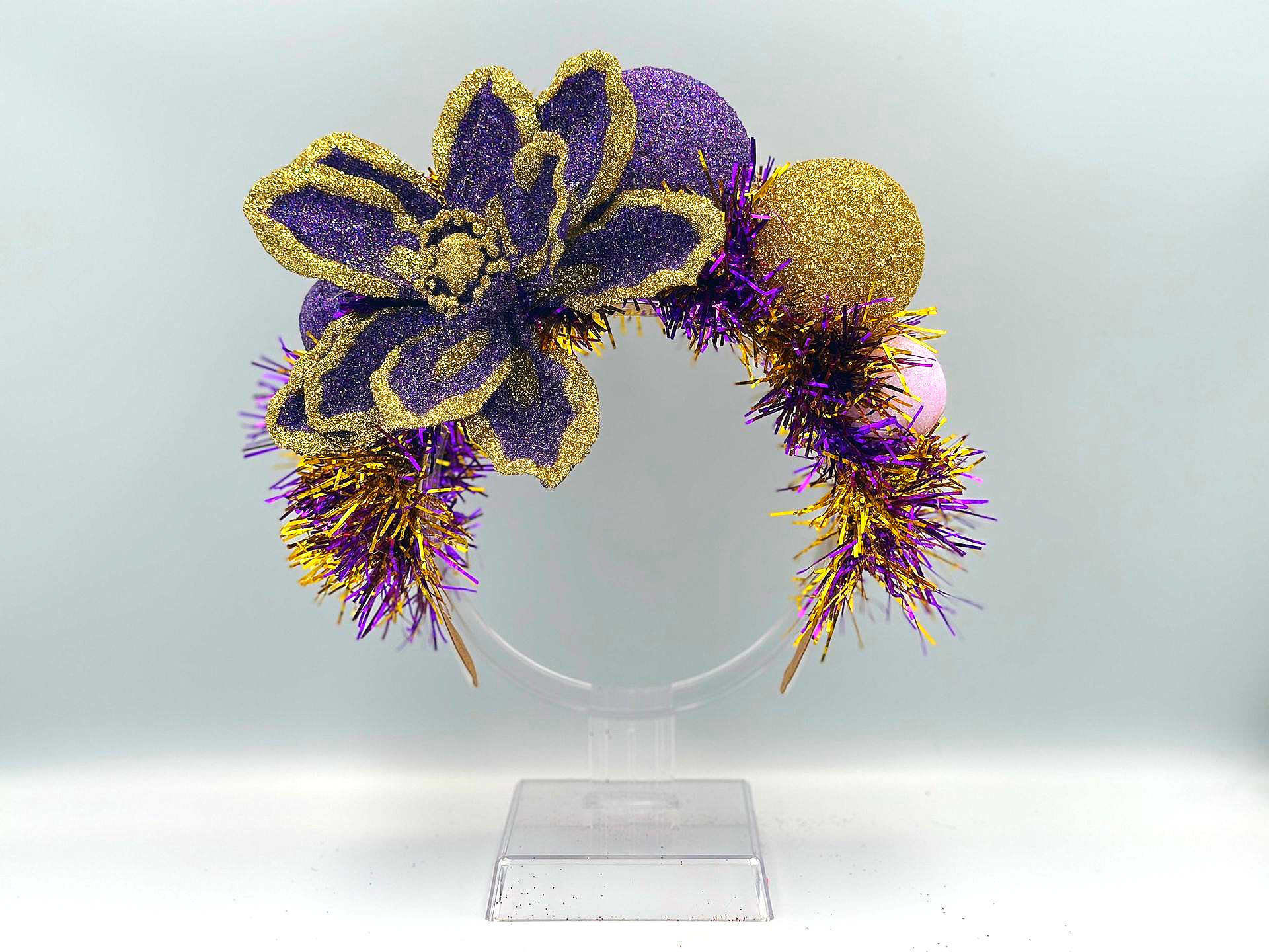 A Mardi Gras themed headband featuring a large glittery purple flower, gold and purple tinsel, and decorative spheres.