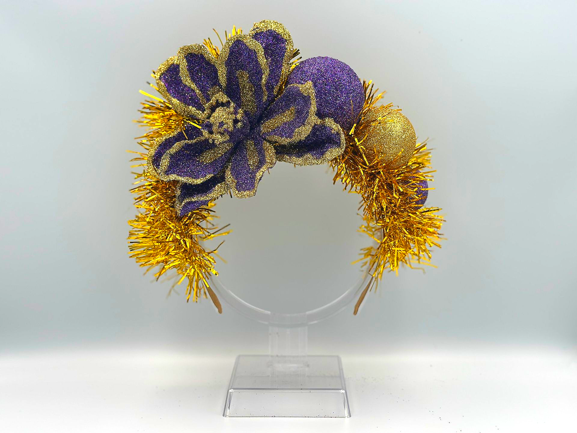 A headband decorated with gold tinsel, a large purple glittery flower, and gold glittery spheres, displayed on a stand.
