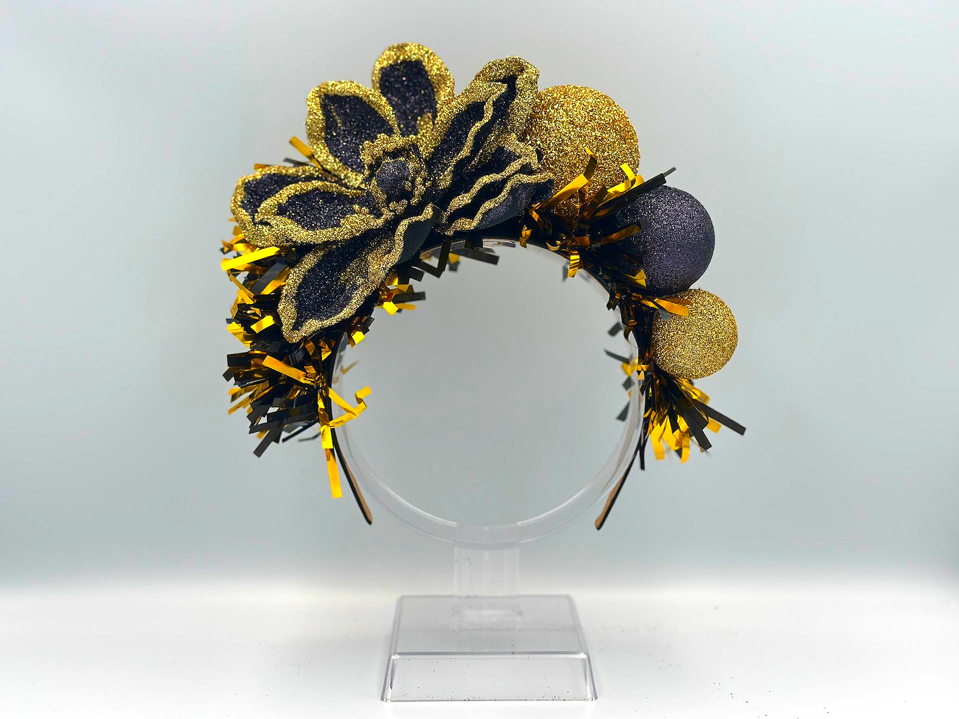 A decorative headband with a large black and gold glitter flower, gold and black baubles, and gold tinsel on a clear stand.