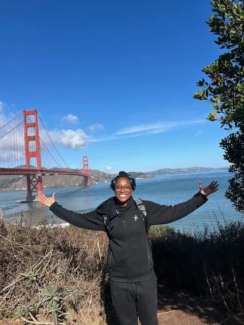 A woman is standing in front of the golden gate bridge with her arms outstretched.