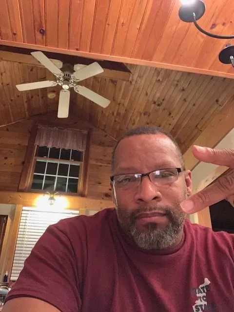 A man wearing glasses and a maroon shirt is making a peace sign